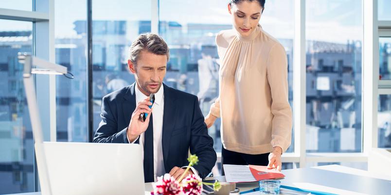 A man in a business suit sits at an office desk with a laptop, holding a voice recorder while a woman standing next to him hands over a red folder