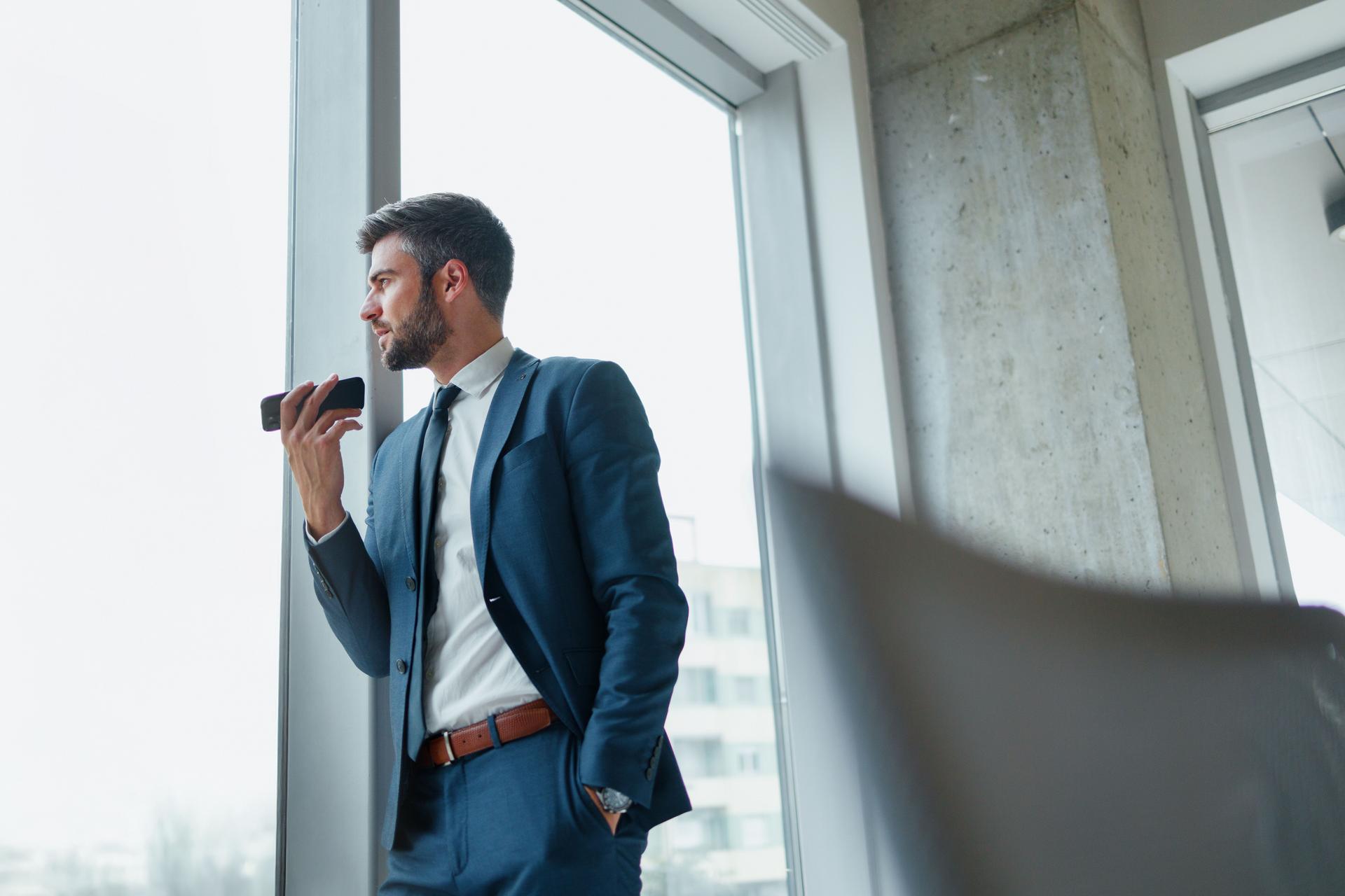 A businessman recording on the phone, by holding it in front of his mouth. He is in the modern corporate building.