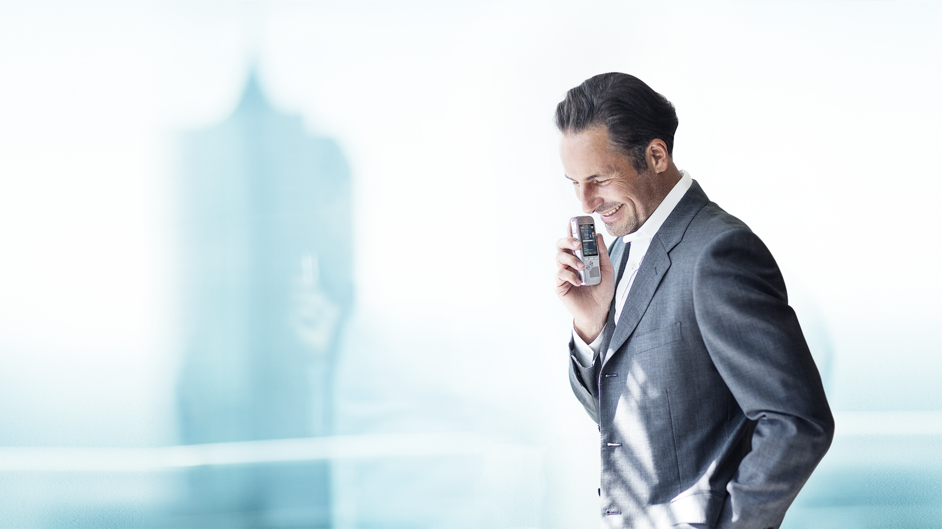 A man in a business suit speaking into a handheld voice recorder, standing in a bright environment.
