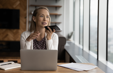 Happy business woman recording audio message on cellphone at workplace, making phone call on speaker, giving audio command to virtual assistant for online search, using voice recognition app