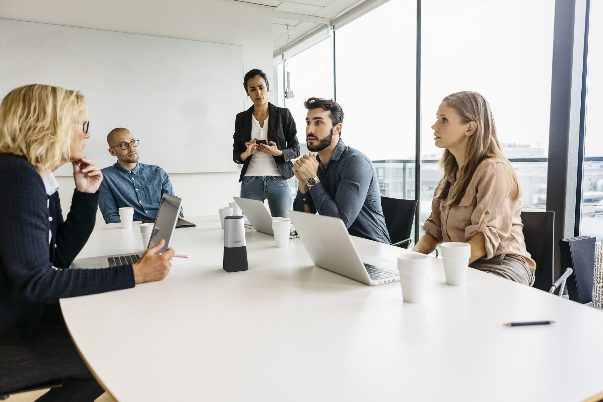 A group of professionals sit around a conference table in a modern meeting room, discussing while laptops are open and a meeting recorder is placed at the center of the table.