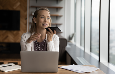 Female read person with long blond hair and white cardigan talking into a phone while using a laptop