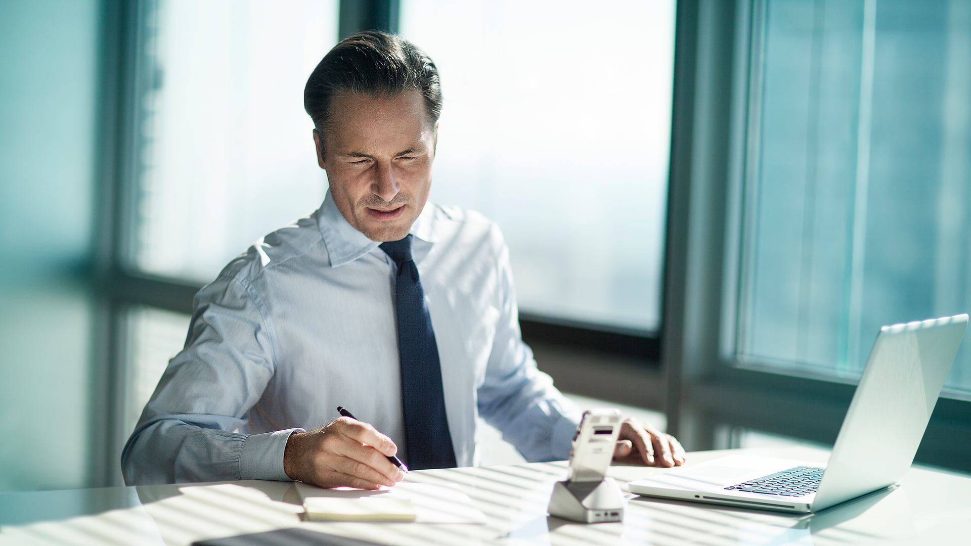 A man in a shirt and tie is sitting at a desk, reviewing notes while his voice recorder sits in a docking station next to his laptop.
