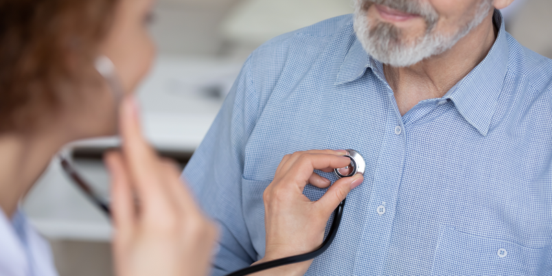 Close up young female general practitioner or cardiologist using stethoscope, checking heartbeat or listening lungs sounds of middle aged elder retired male patient at checkup appointment in hospital.